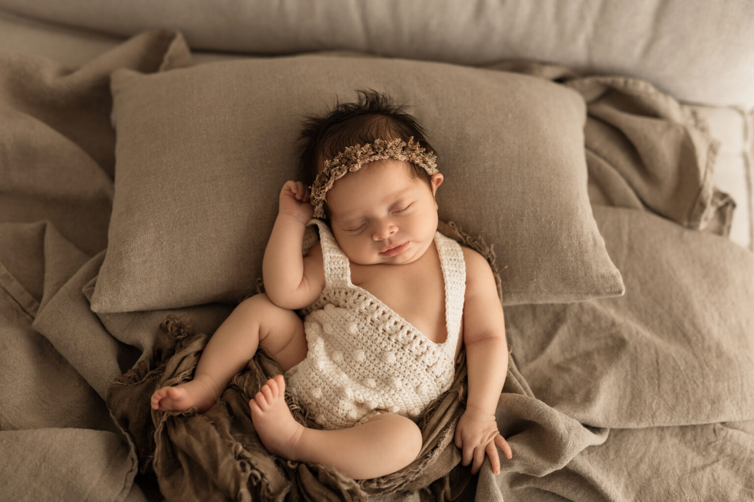 525A8254 Newborn girl peacefully sleeping on neutral linens, wearing a handmade crochet romper and a delicate floral headband for a soft, natural look.